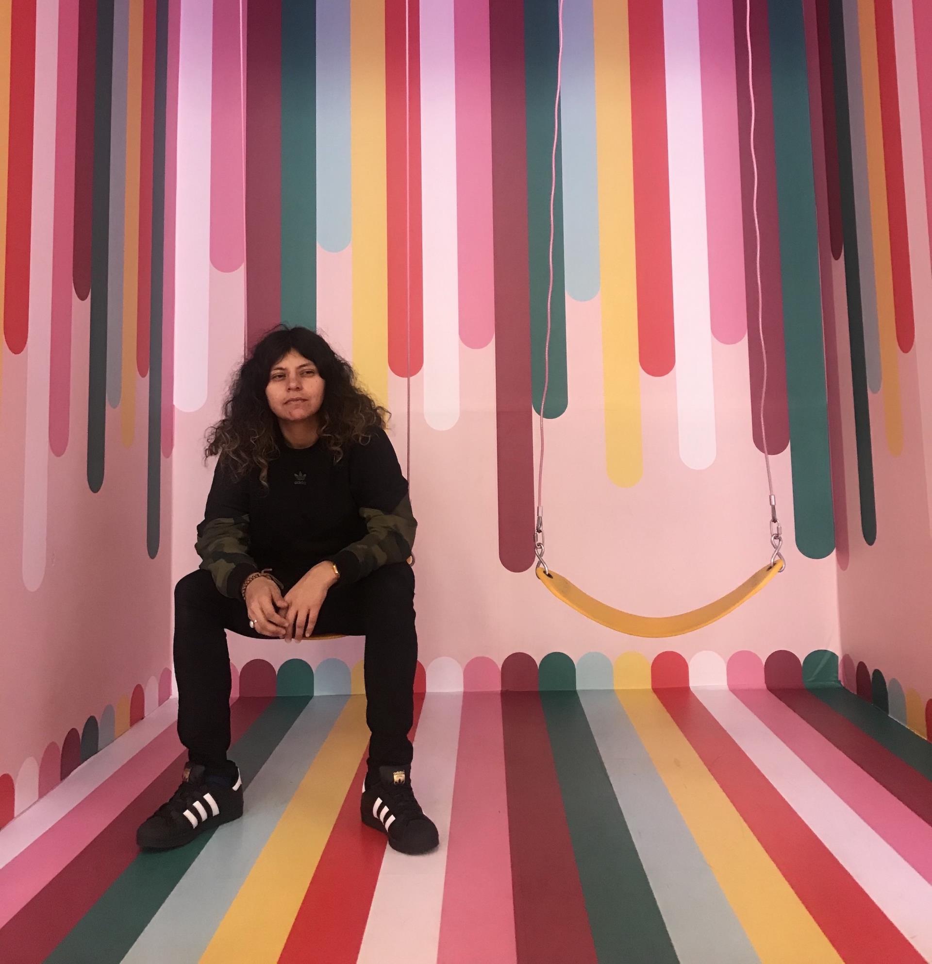 Artist Ruby Barrientos sitting on a swing in a small room painted with colorful stripes.