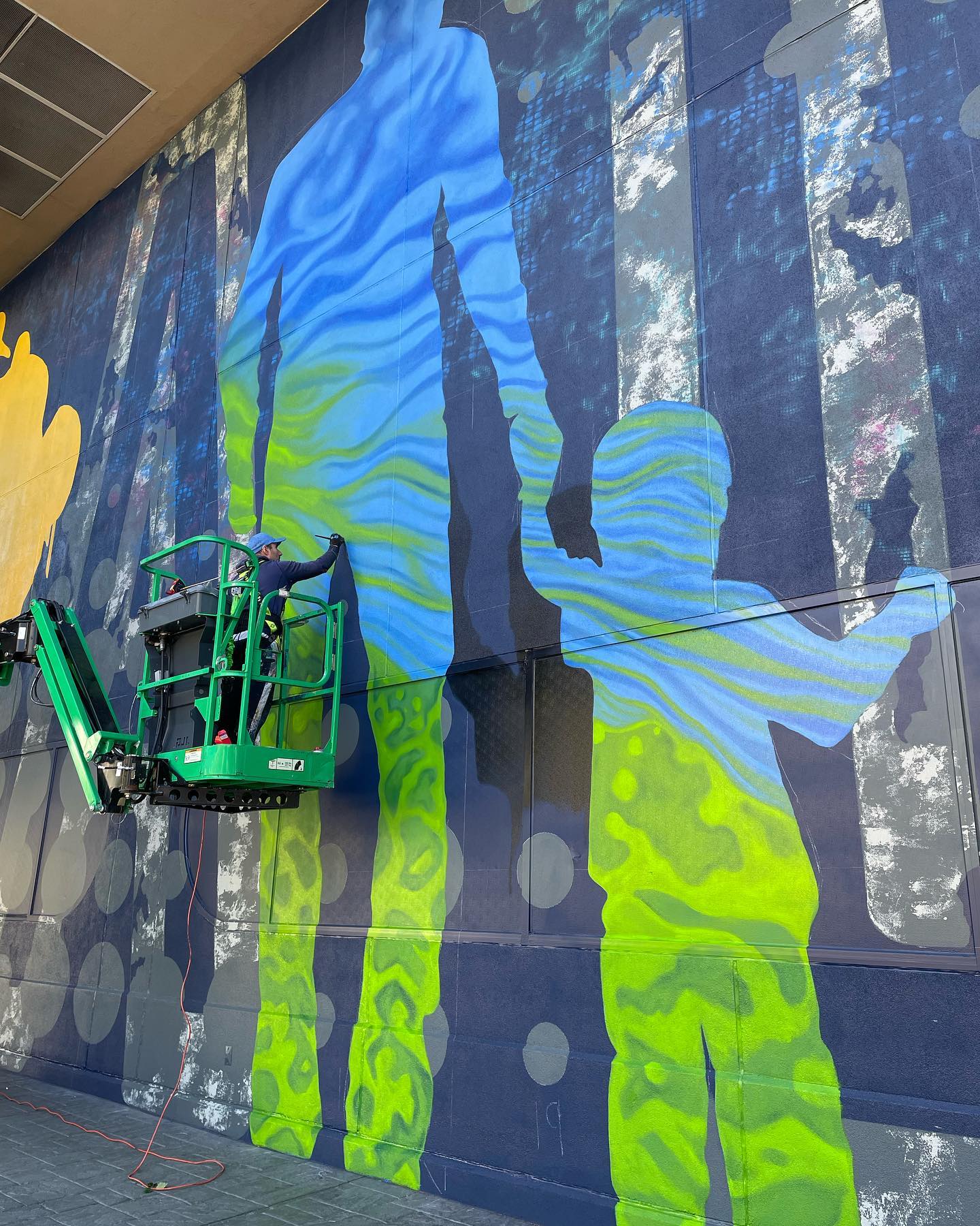 Artist Rafael Blanco is on a lift painting a large mural on the side of the National Bowling Stadium